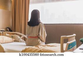 Back view of patient woman sitting on bed in hospital ward looking out at window.