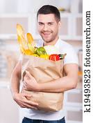 He is passionate about healthy food. Happy young man holding shopping bag full of groceries while standing in the kitchen