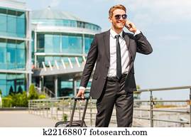 Executive on the Go. Handsome young businessman in full suit carrying suitcase and talking on the mobile phone while walking outdoors