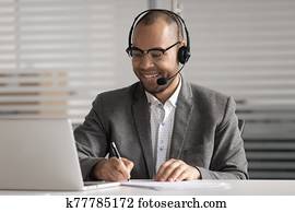 Happy African American employee in headset making notes, using laptop Happy African American employee in headset making notes, using laptop