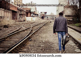 Young man with guitar case is going away among industrial ruins