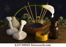 An arrangement of turmeric, spice, oil bowl and bottle, and massage poultice boluses used in Ayurveda massage, with an exotic flower and incense burning in the background.