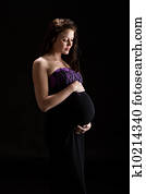 Beautiful brunette model in last trimester posing in studio in full length dress on black background looking lovingly at baby bump