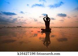 Yoga woman sitting in lotus pose on the beach during sunset, with reflection in water.
