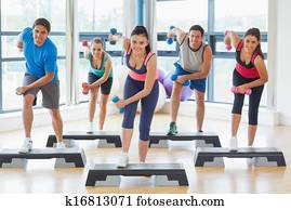 Full length of instructor with fitness class performing step aerobics exercise with dumbbells in a gym Full length of instructor with fitness class performing step aerobics exercise with dumbbells in a gym