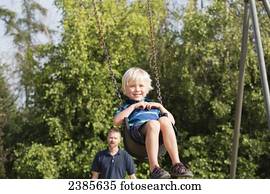 A boy swinging on a swing set with his father in the background; Stony Plain, Alberta, Canada