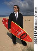 Man Standing On Beach In Business Suit Holding A Surfboard.