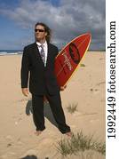 Man Standing On Beach In Business Suit Holding A Surfboard.