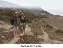 A young couple stands on a rugged dirt trail on a mountainous landscape looking out; Praia da Cordoama, Portugal