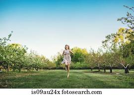 Full length front view of young woman wearing sleeveless dress running through orchard, looking at camera smiling