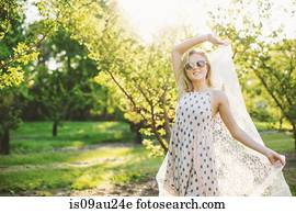 Young woman in orchard wearing sleeveless dress and sunglasses holding lace fabric, looking at camera smiling