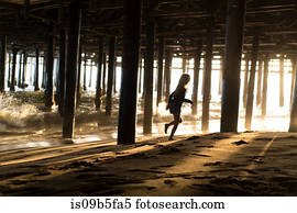 Silhouette of girl running from lapping waves underneath Santa Monica pier, California, USA