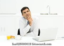 Businessman sitting at table with laptop computer and breakfast, hand under chin, looking up
