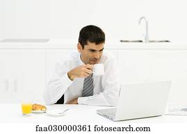 Businessman sitting at kitchen table, looking at laptop computer, drinking coffee