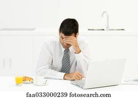 Businessman sitting at breakfast table with laptop computer, holding head