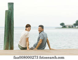 Couple sitting on edge of dock, smiling over shoulders at camera