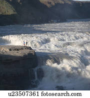 Whitewater flowing into waterfall in valley past rock outcrop with sightseers