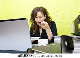 Caucasian young woman eating and working on her laptop computer at her desk