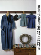 TEXTILES: An Amish maternity dress and antique children's clothing hang on a peg rack , 1844 coverlet folded on dresser below, grapevine wreath