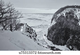 Monochrome aerial view of floating sea ice in a channel : deep gully with steep cliffs either side in the snow, lighthouse on top, with open sea beyond, Shiretoko National Park, Hokkaido, Japan
