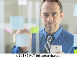 Businessman with smartphone sticking notes on office glass wall