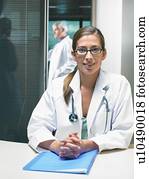 Portrait of female doctor at her desk with make doctor in background.