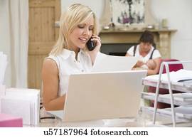 Woman in home office with laptop and telephone with mother and baby in background