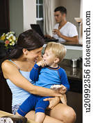 Caucasian woman with toddler son in kitchen with father on laptop in background.