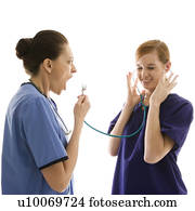 Portrait of Caucasian healthcare workers wearing scrubs with one yelling into stethoscope that is attached to other's ears against white background.