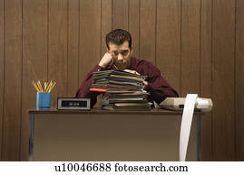 Caucasion mid-adult retro businessman with bored expression sitting at desk piled high with folders.